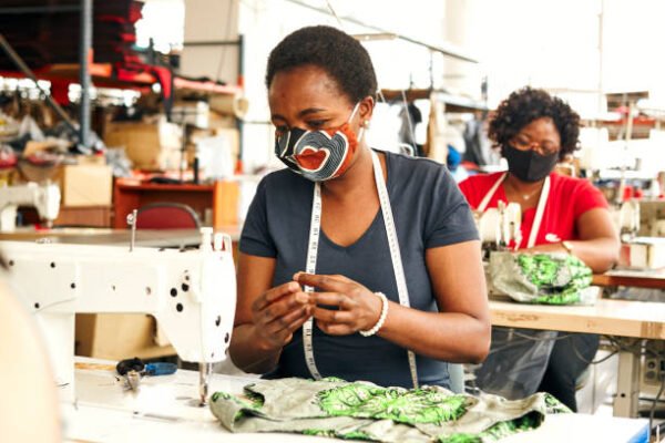 Two black African Zimbabwean ladies wearing coronavirus face masks working in a textile factory on the sewing line using sewing machines, measuring tapes and scissors to stitch lining in African styled patterned fabric garment